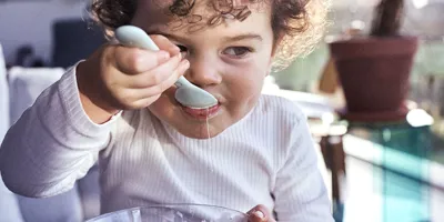 Un bambino con i capelli ricci e scuri mangia lo yogurt con un cucchiaio da una piccola ciotola di vetro.
